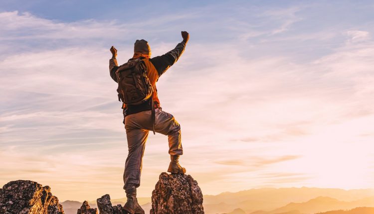 hiker with backpack at the top of a rock with his hands raised e