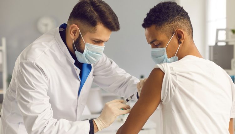 Young doctor in face mask administering Covid-19 or flu antiviral vaccine to patient