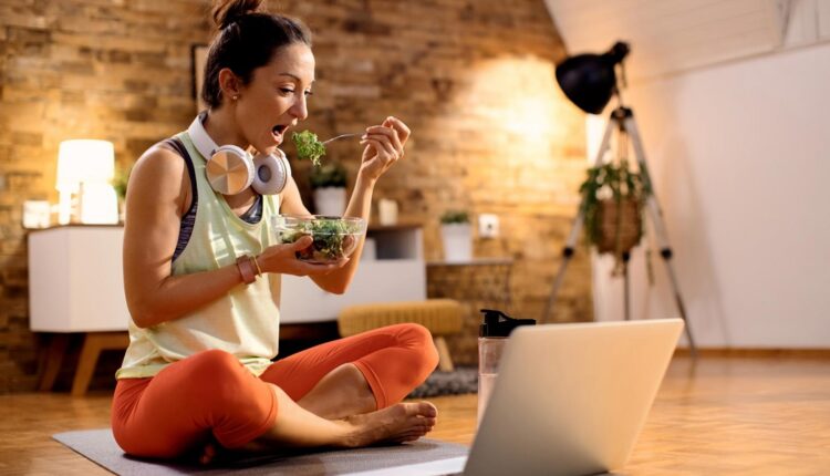 female-athlete-eating-salad-using-laptop-after-working-out-home